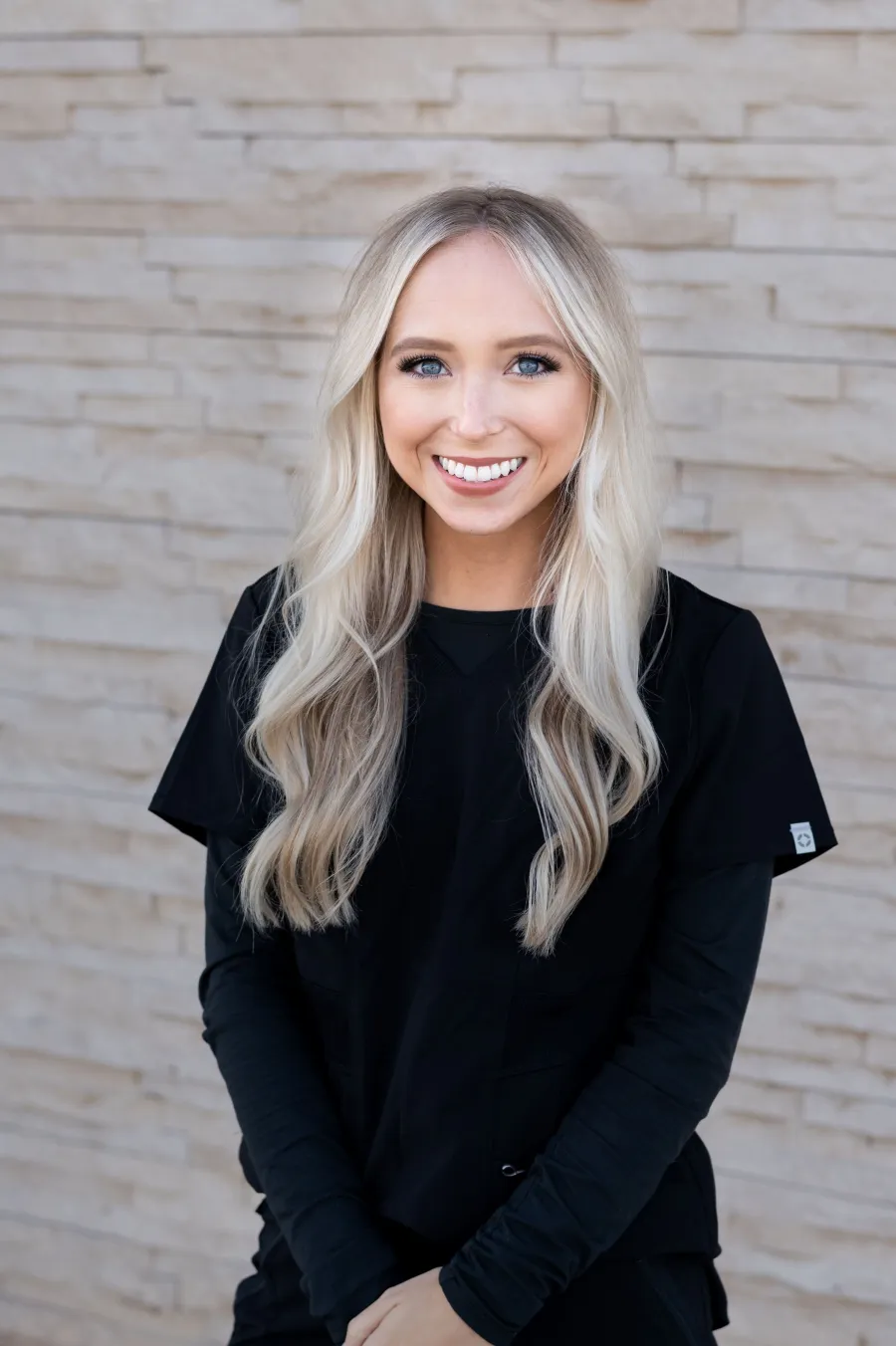 Jesica Jordan of Dahlia by WHA, smiling, in black scrubs against a pale stone wall